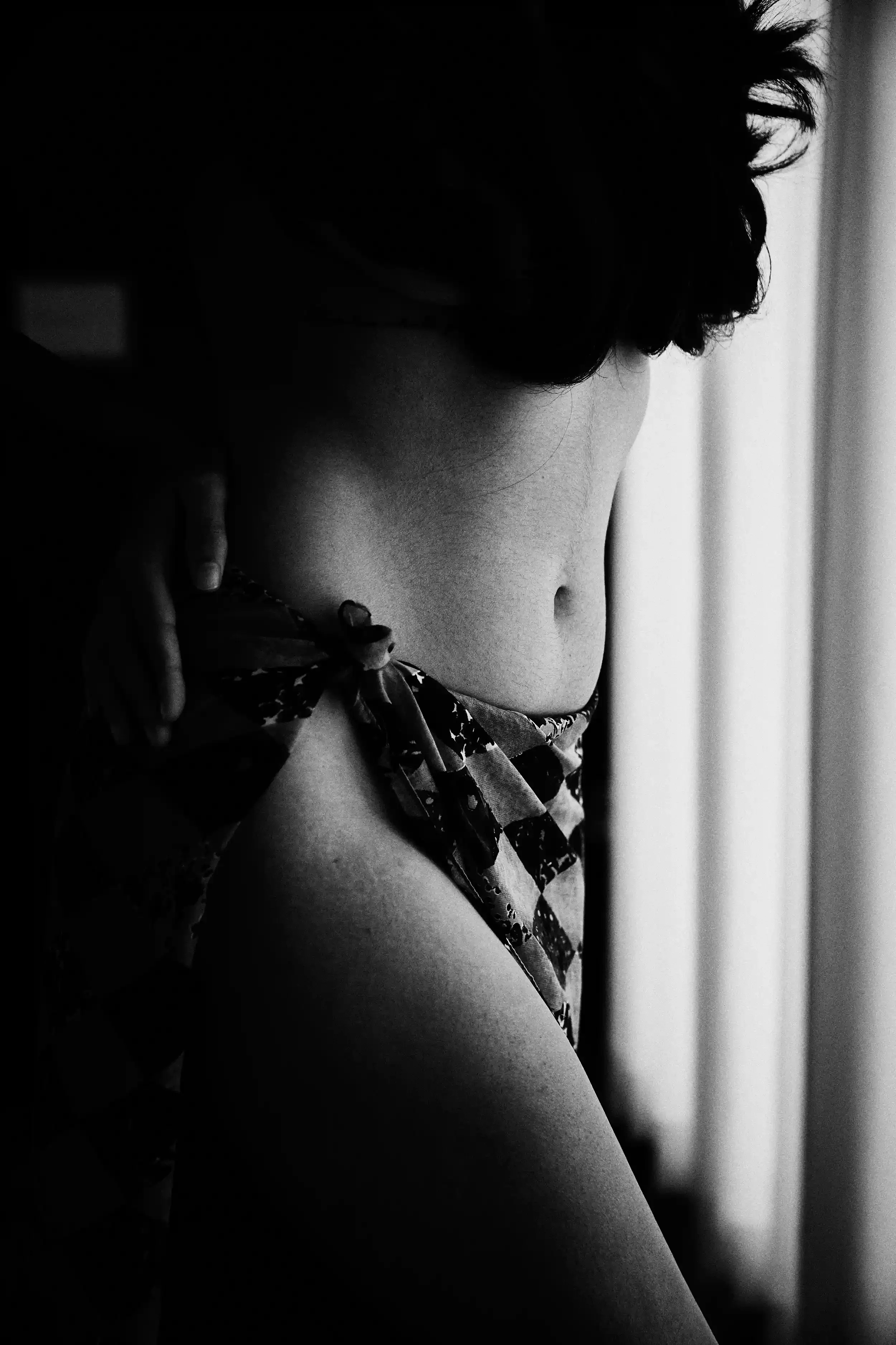 Black and white close-up photograph of a woman with short, curly hair, shown from the side, wearing a patterned top, with her face and body partially turned away from the camera, standing near a light source on the right side of the image. anandclix.