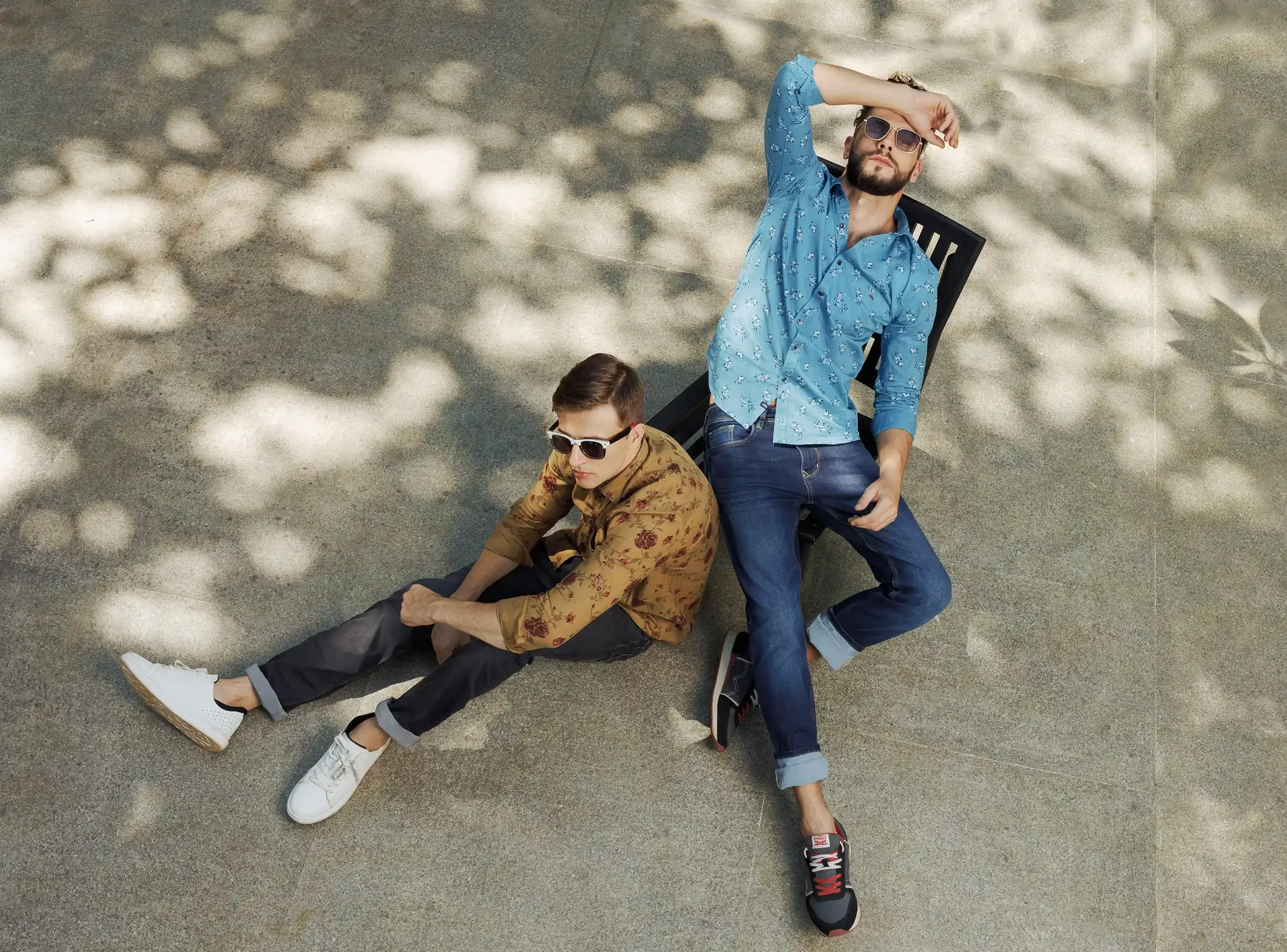 Two men wearing sunglasses are sitting and lying on the ground, with one leaning back against a chair, on a concrete surface with dappled sunlight. Anandclix, Bangalore, advertising photographer, Fashion photographer, Editorial.