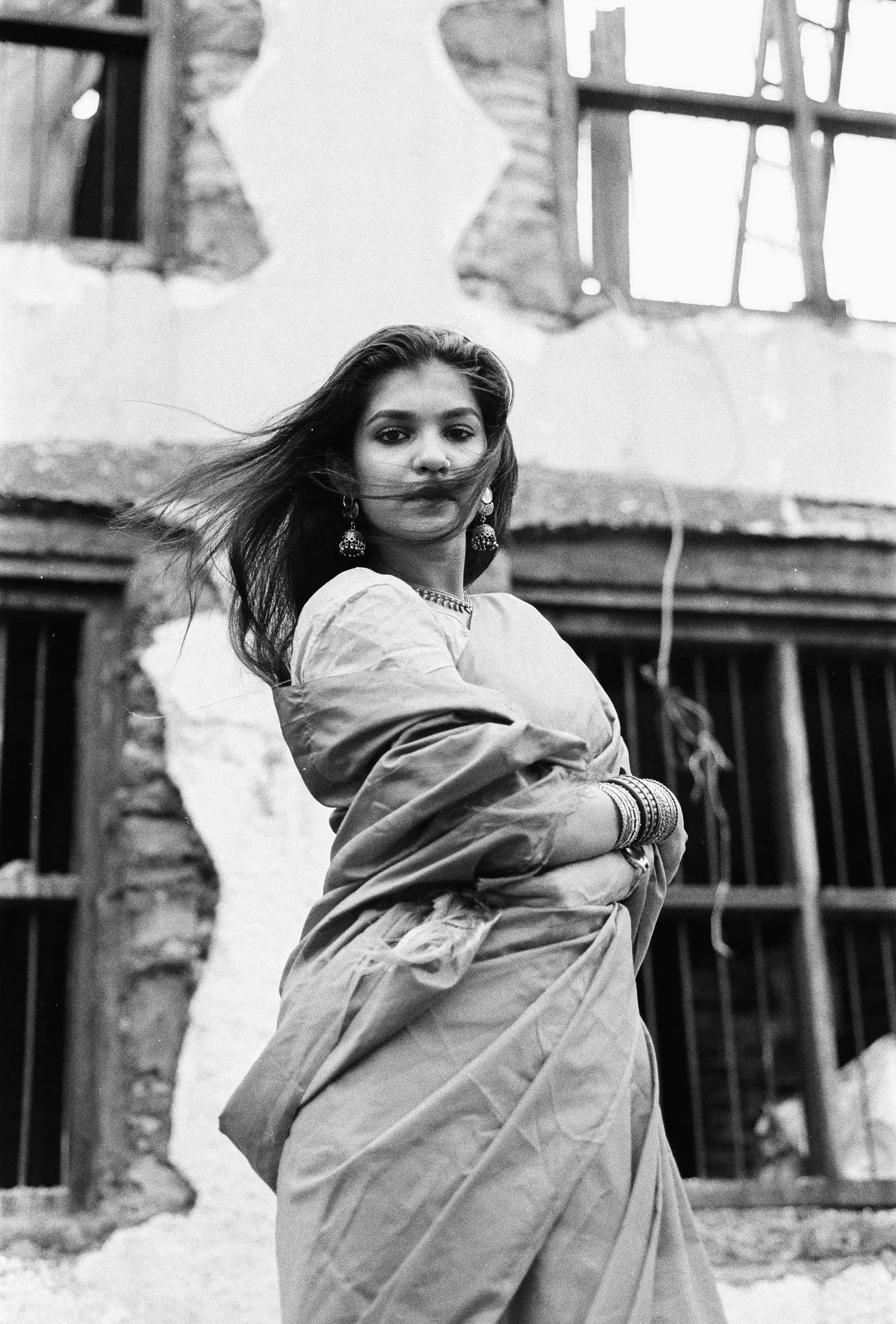 A woman wearing traditional Indian attire, jewellery, and earrings, standing in front of a building with broken windows and an exposed brick wall. Film Photography, Analogue photography, Anandclix, Fine-art, Fine-art portrait.