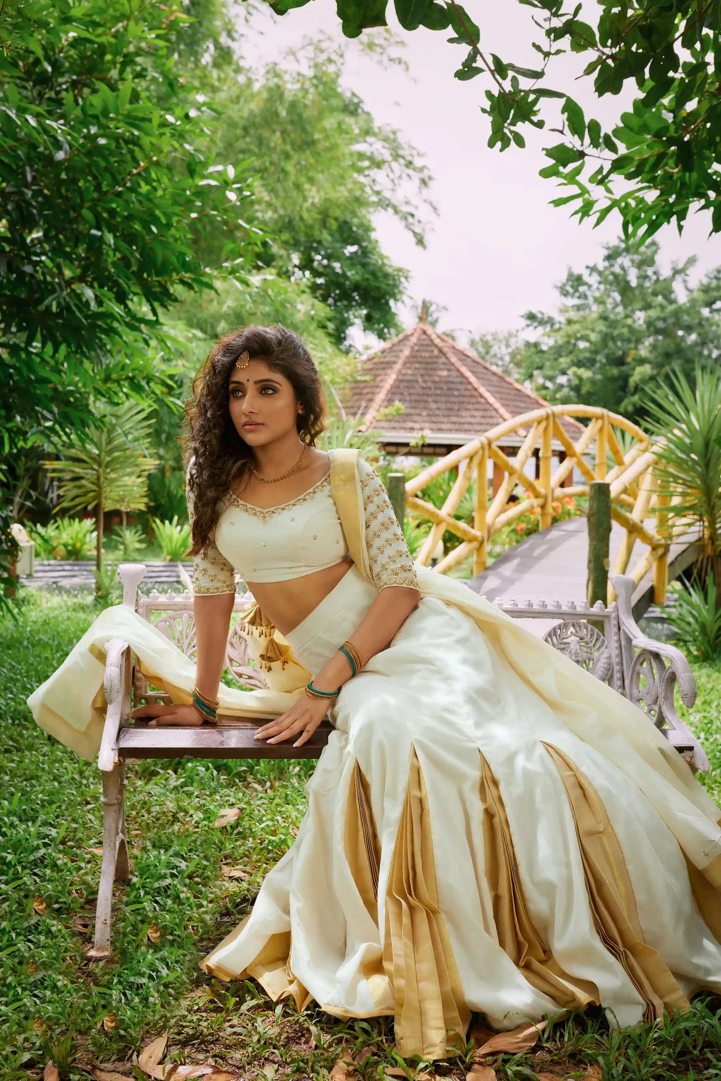 A woman dressed in traditional Indian attire, sitting on a wooden park bench surrounded by greenery and plants, with a small bridge and a gazebo in the background.