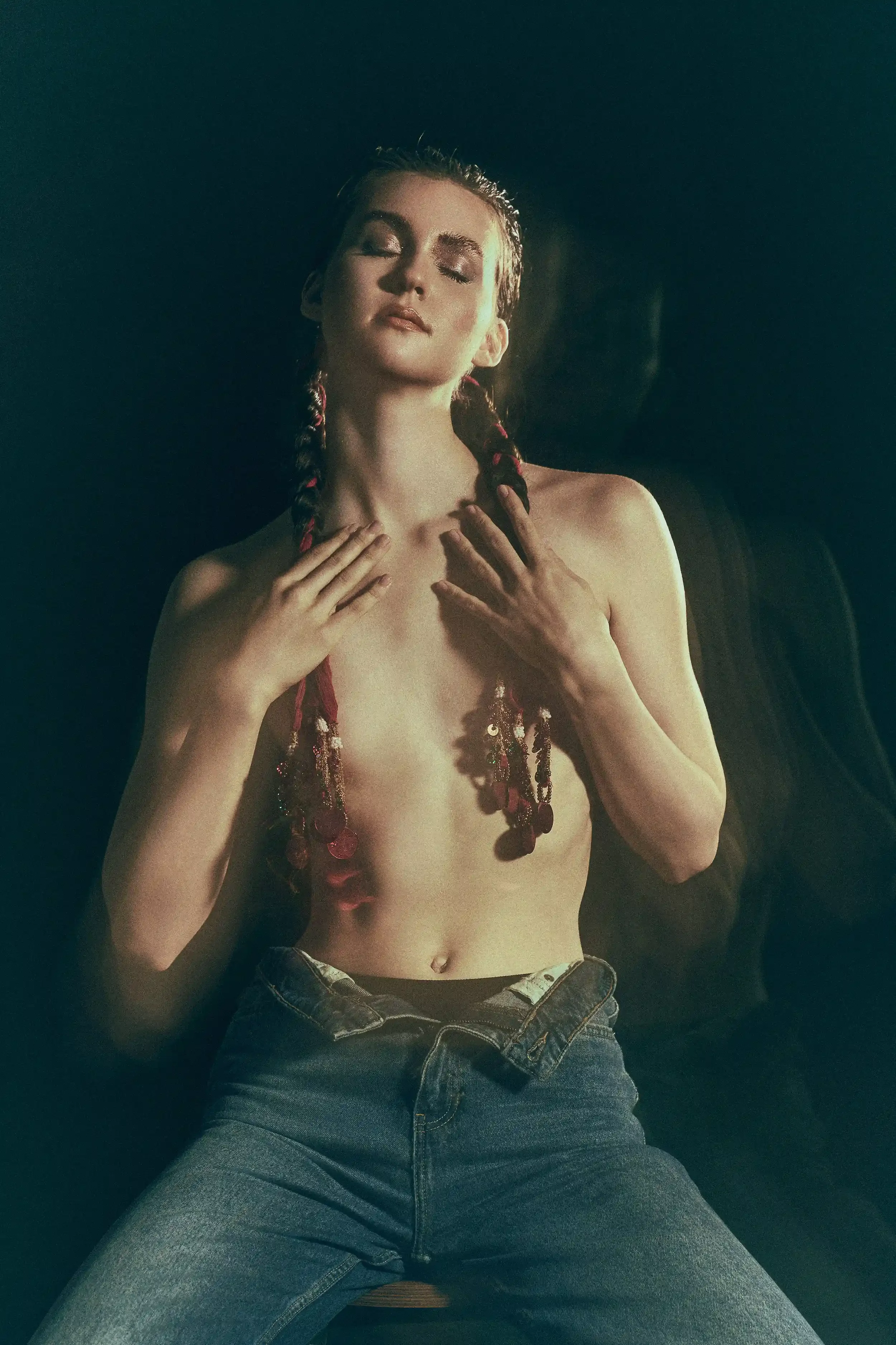 A topless woman with braided hair and pink highlights, wearing jeans, in a dark setting, posing with eyes closed and hands touching her neck and chest.