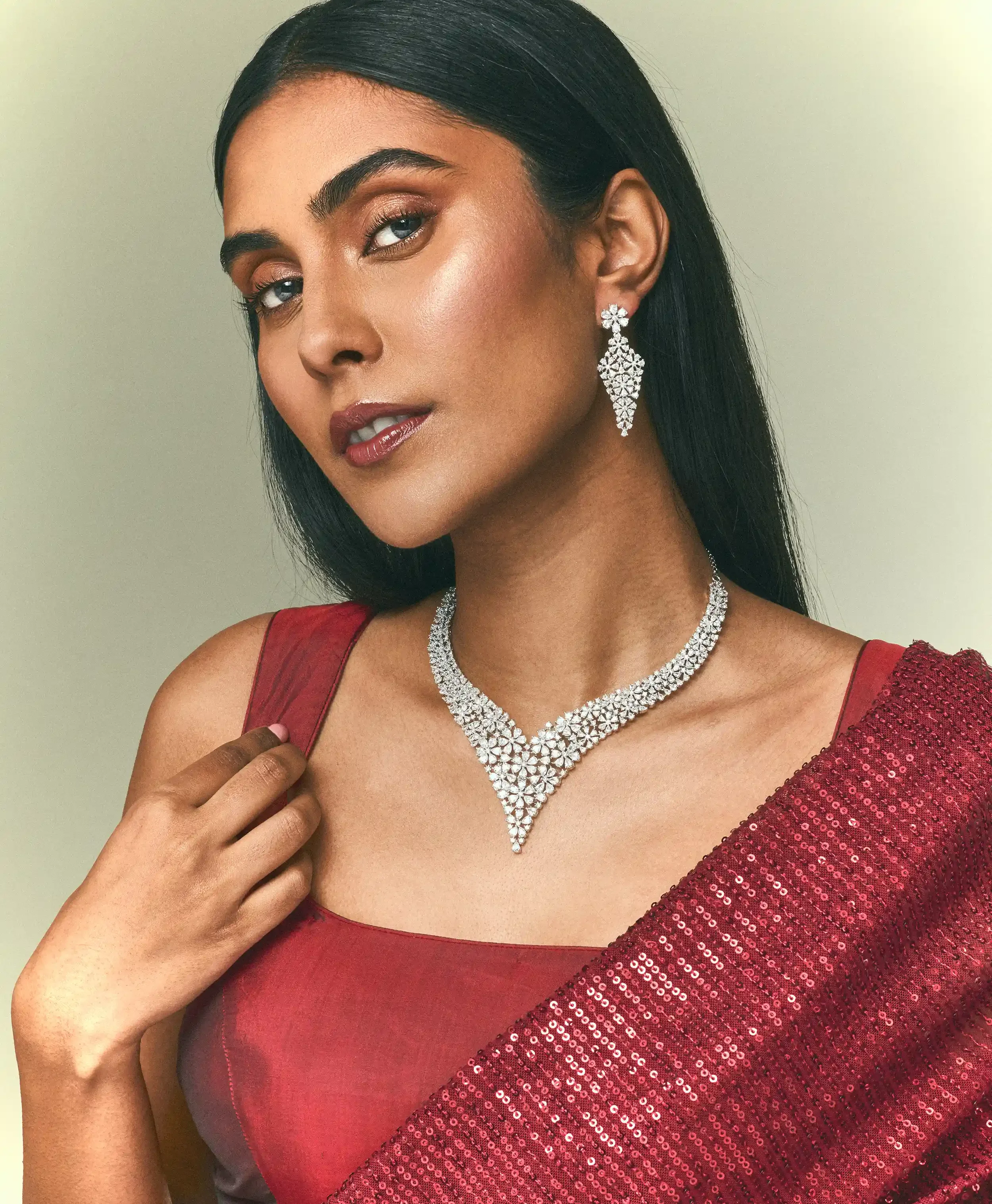 A woman wearing a red sequined dress, diamond jewellery, including a necklace and earrings, long dark hair, and makeup, holding her dress strap with one hand against a plain background. Kushal's Fashion Jewellery, Anandclix, Bangalore, advertising.