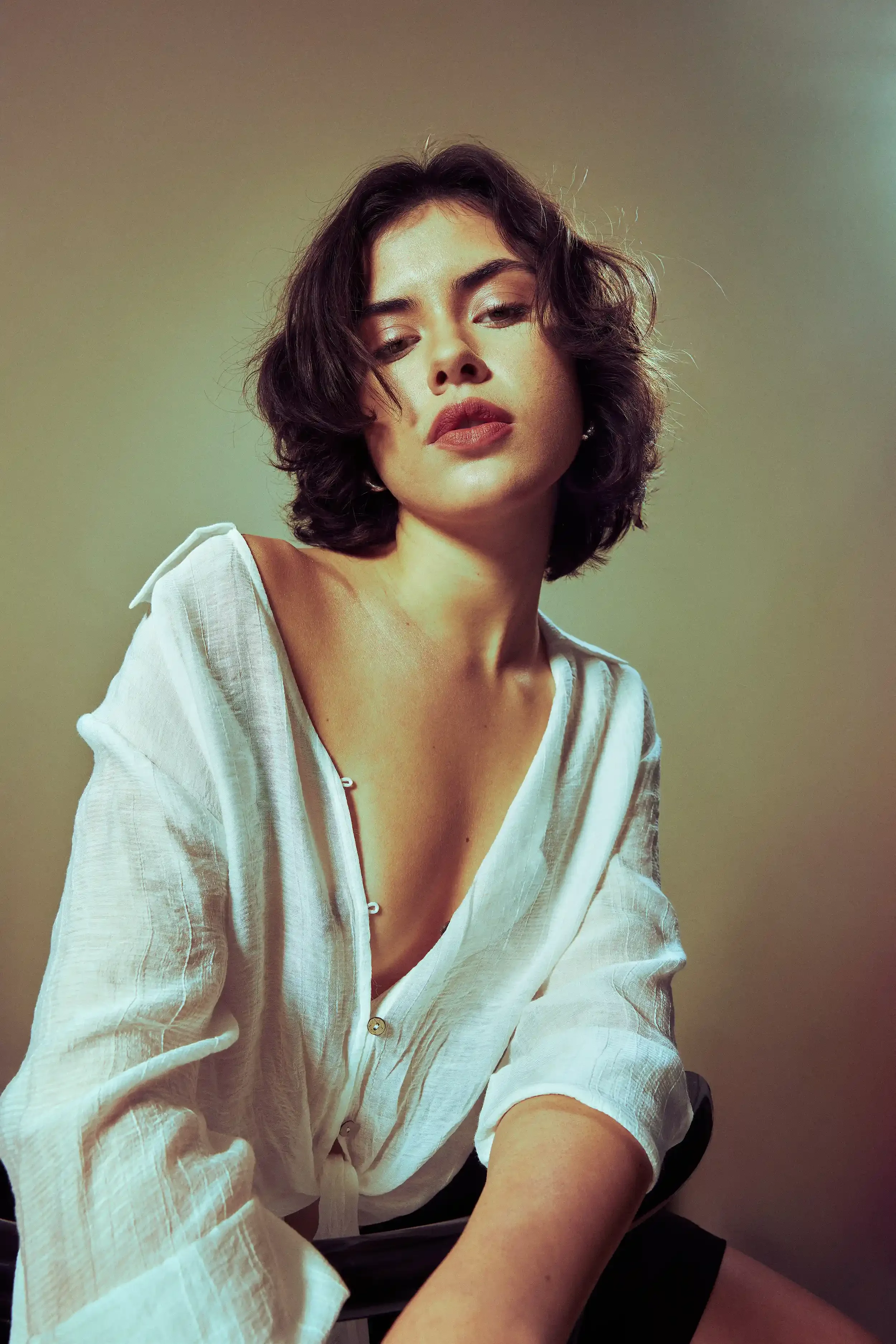 A woman with dark, wavy shoulder-length hair, wearing a white button-up shirt with the top few buttons undone, looking directly at the camera with a serious expression against a neutral background.