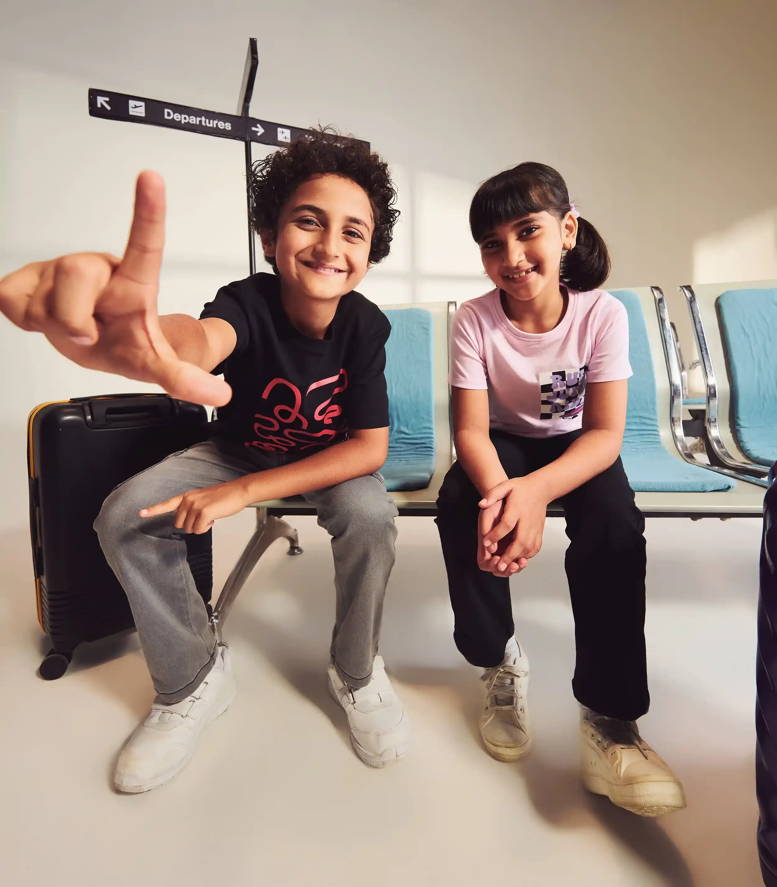 Two children sitting on blue chairs at an airport, smiling at the camera. The boy on the left has curly hair, wears a black t-shirt, gray pants, and white sneakers, and is pointing towards the camera with his finger. The girl on the right has straigh