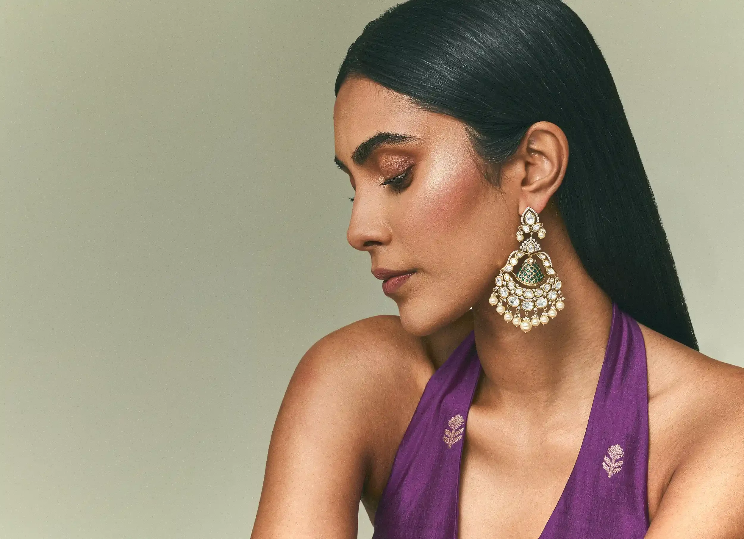A woman with long black hair wearing ornate pearl and gemstone chandelier earrings and a purple halter top with small floral embroidery, looking downward against a plain light green background. Anandclix, Bangalore, advertising photographer, Fashion.