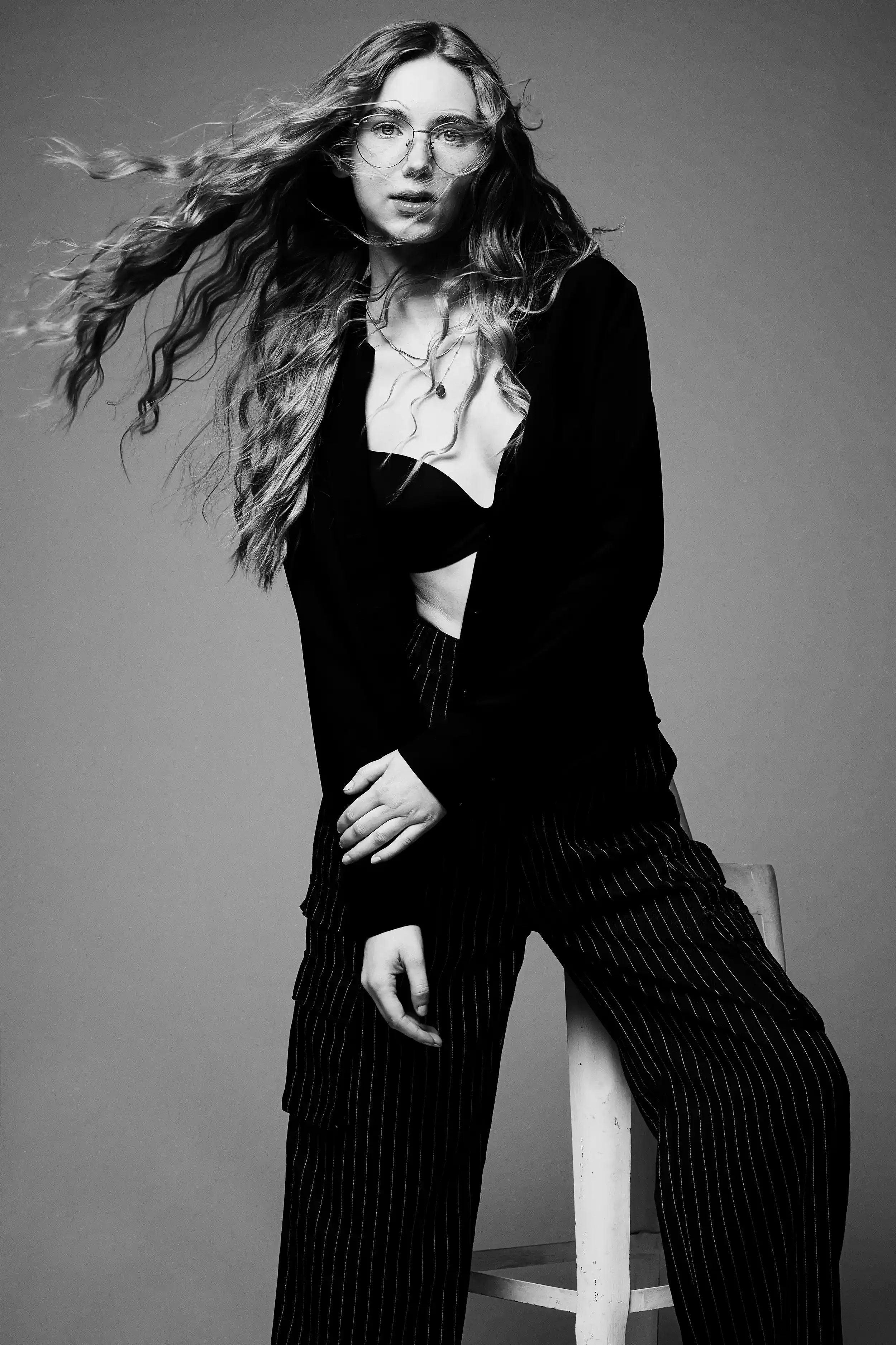 A black-and-white photo of a woman with long, wavy hair wearing glasses, a black blazer, a bra top, and pinstriped pants. She is sitting on a chair with her legs apart, one hand resting on her lap, and the other holding the chair. Her hair is blowing
