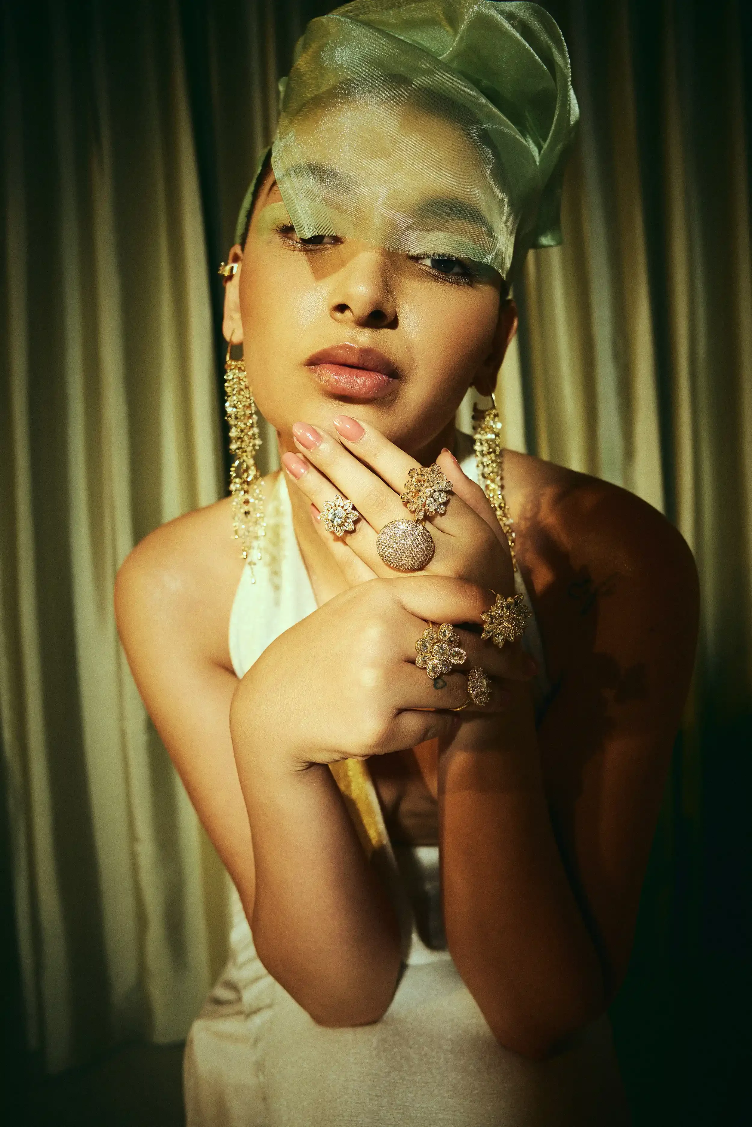 A woman with gold jewellery, including earrings and rings, poses with her hand near her face, wearing a head covering and a satin dress, with a curtain background. Anandclix, Bangalore, advertising photographer, Fashion photographer, Editorial.