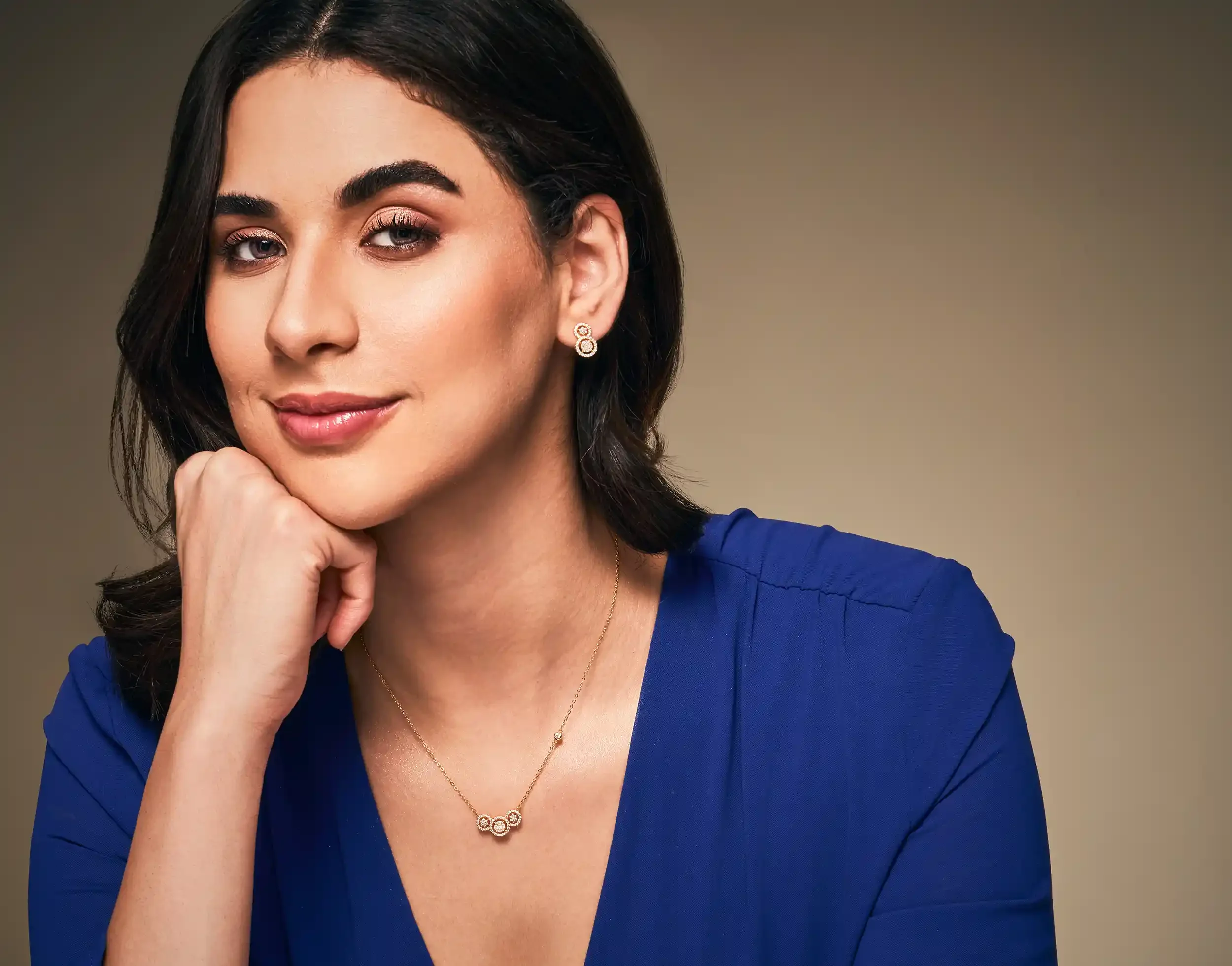 A woman with dark hair and fair skin, wearing a blue dress and jewellery, including earrings, a necklace, and a ring, rests her chin on her hand and smiles gently. Kushal's Fashion Jewellery, Anandclix, Bangalore, advertising photographer, Fashion.