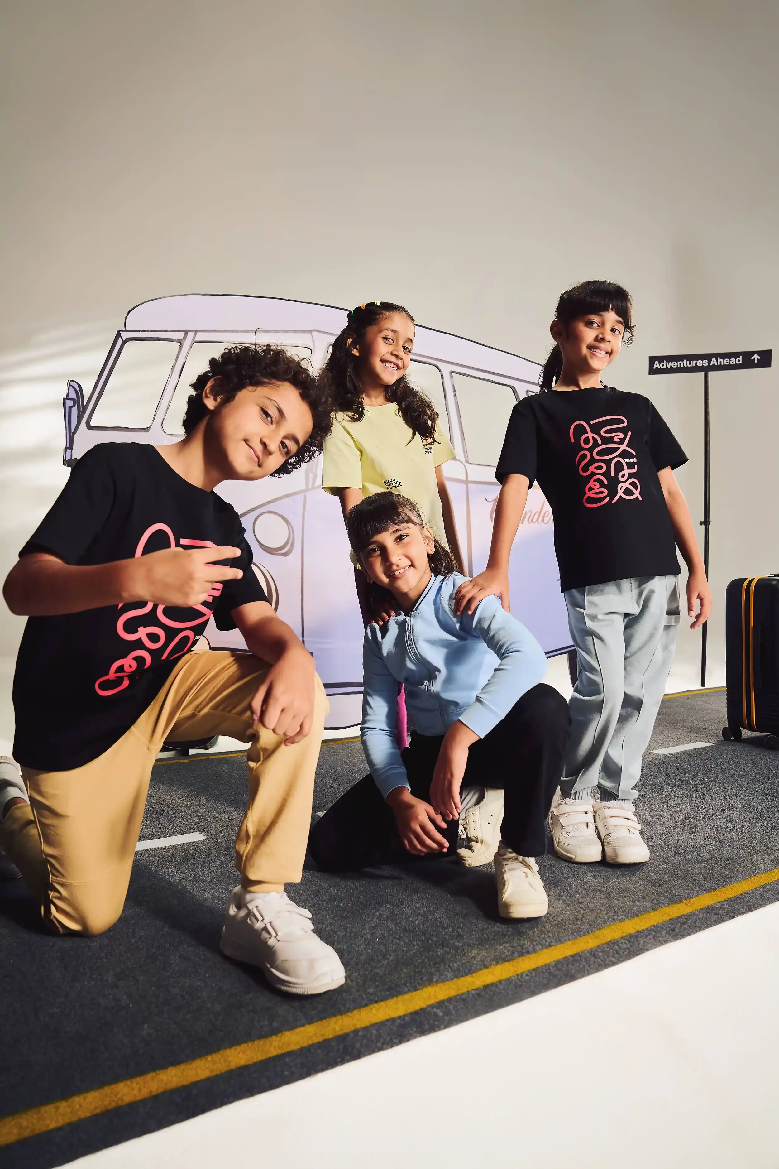 Four children posing in front of a colourful backdrop of a camper van, with a sign that reads "Adventures Ahead" and a suitcase nearby. Anandclix, Bangalore, advertising photographer, Fashion photographer, Editorial photographer.