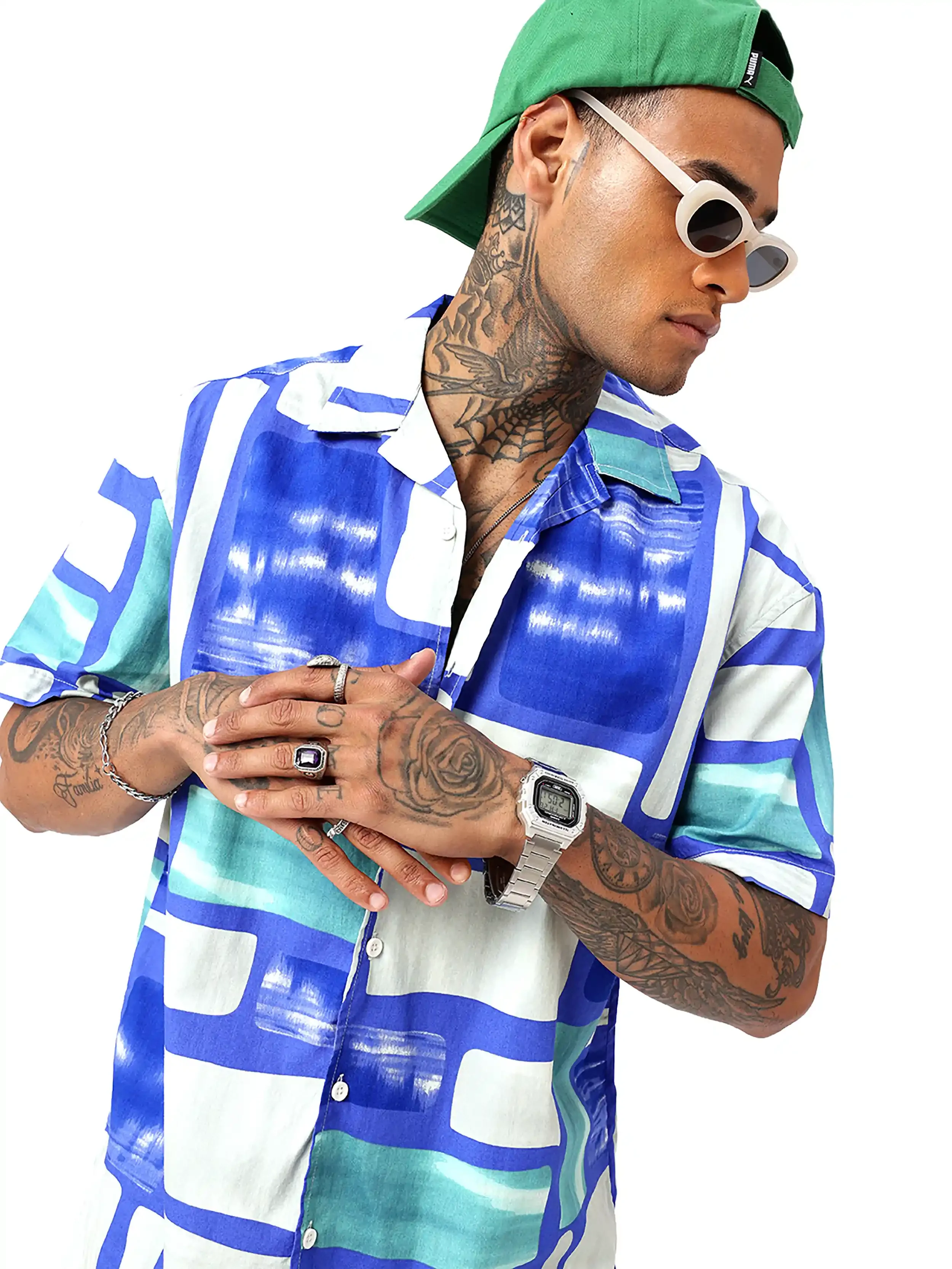 Man wearing white sunglasses, green cap backward, blue and white patterned short sleeve shirt, with tattoos on neck and arms, wearing rings and a digital watch, standing against a plain white background.