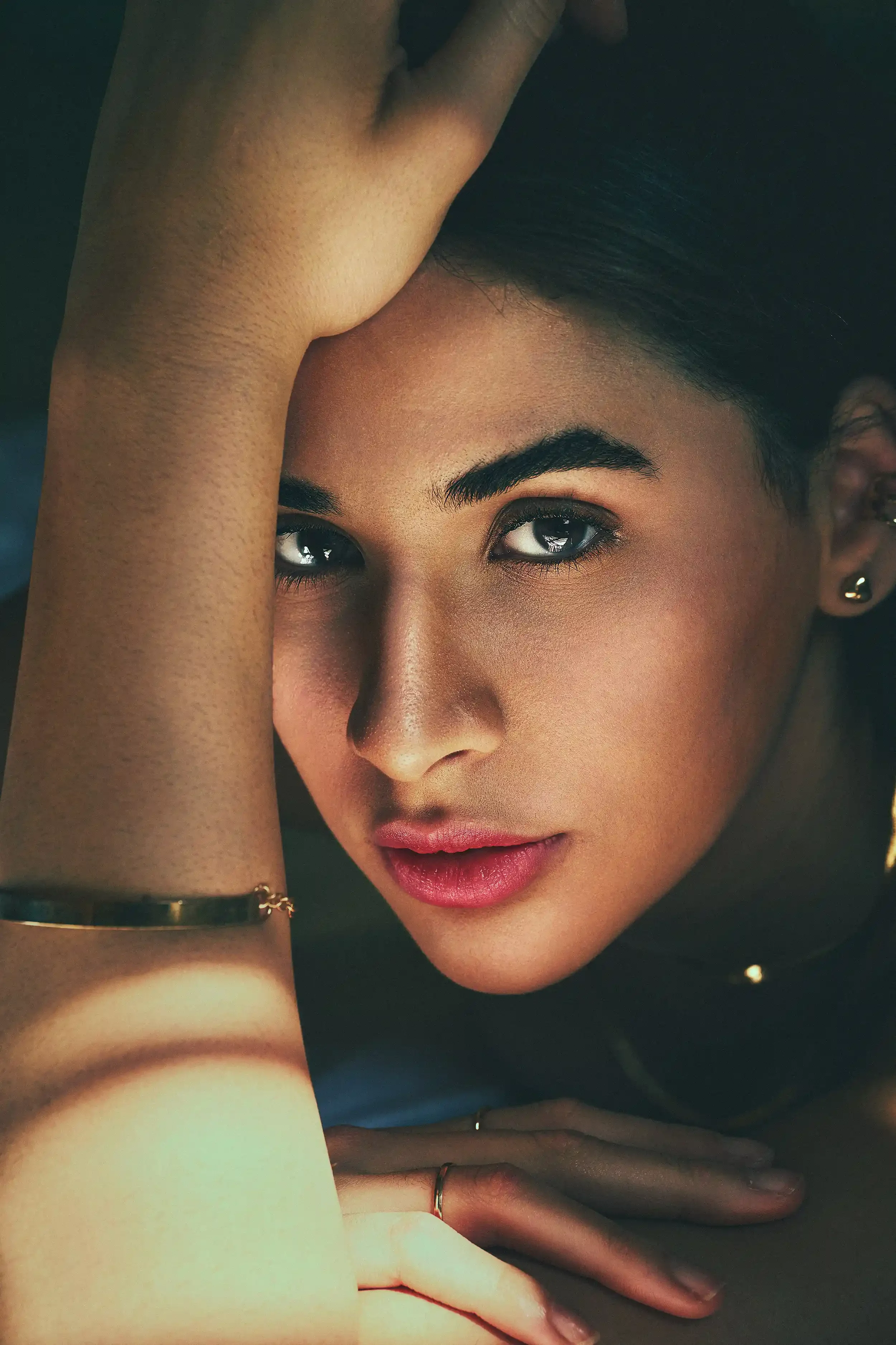 Close-up of a woman with dark hair, blue eyes, and pink lipstick, resting her arm on a surface with her hand near her face, wearing jewelry including a ring, earrings, and a bracelet. Anandclix, Bangalore, advertising photographer, Fashion.