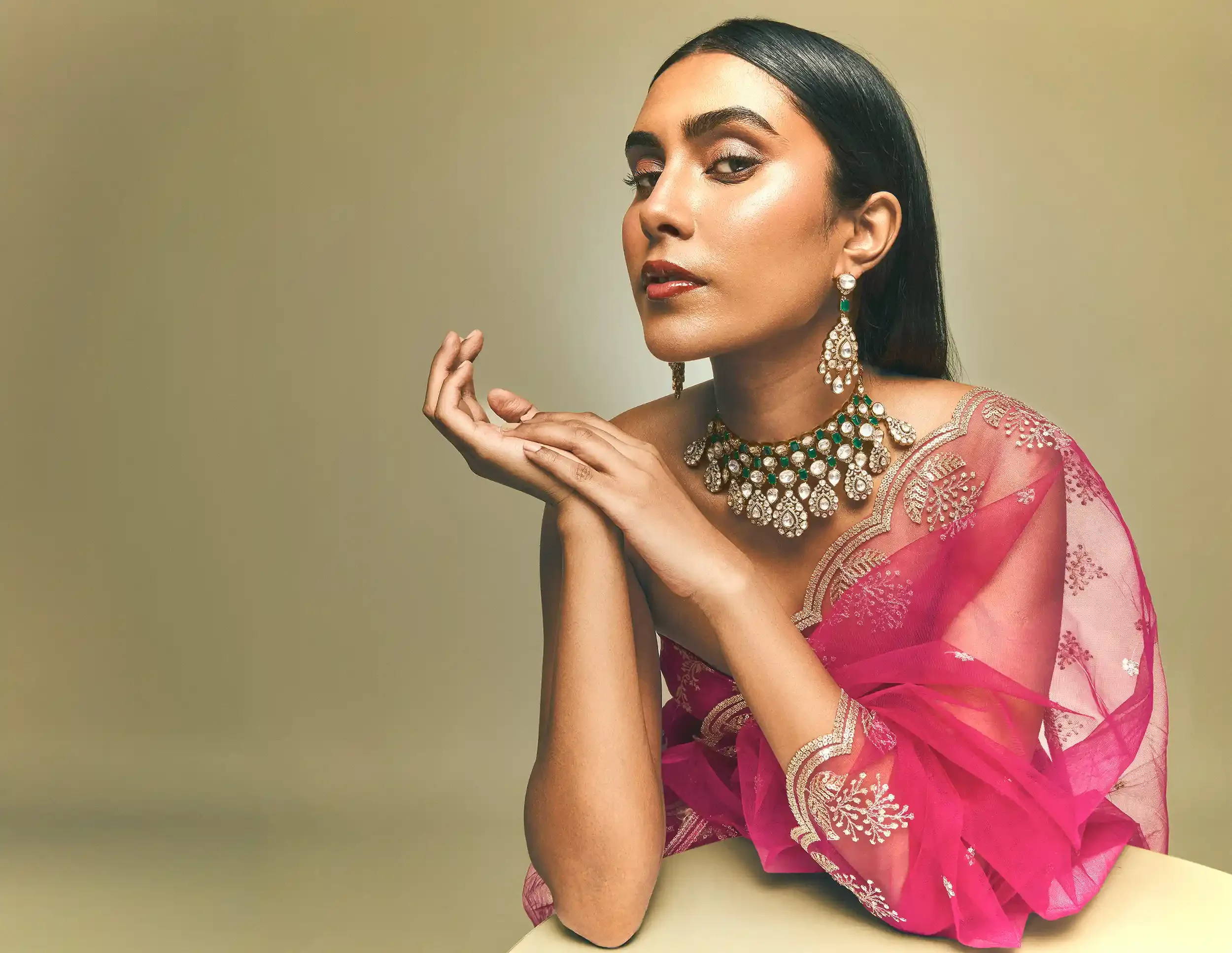 A woman with dark hair styled straight, wearing intricate gold and emerald jewelry, and dressed in a pink saree with gold embroidery. She poses with her head slightly tilted, eyes half-closed, and her hands gently near her face against a plain beige 