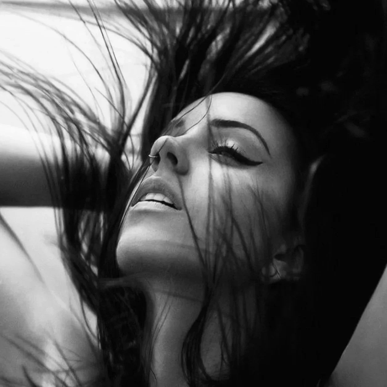 Zwart-wit foto van een vrouw met lange, donkere haar die over haar gezicht ligt, met make-up en een uitgesproken oogmake-up. Ze ligt op haar rug met haar hoofd schuin omhoog.