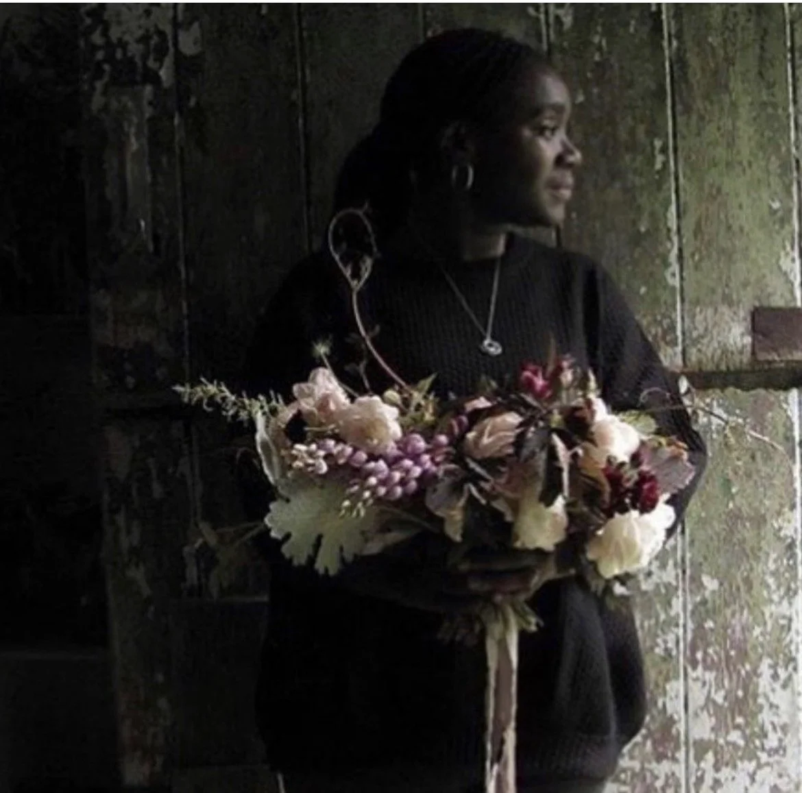Florist Catherine Foxwell holding a bouquet of flowers, standing in a dimly lit space with a weathered wall background.