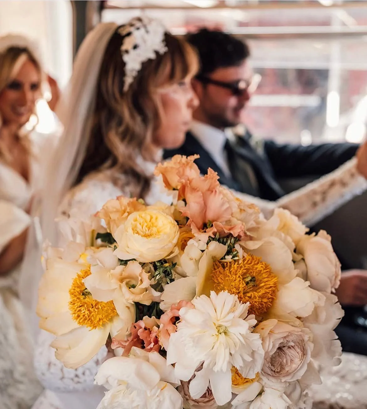 A wedding bouquet with white and peach-colored flowers in the foreground. In the background, a bride in a lace dress and veil, a groom in a tuxedo, and another woman are visible, all sitting and smiling at a wedding ceremony.