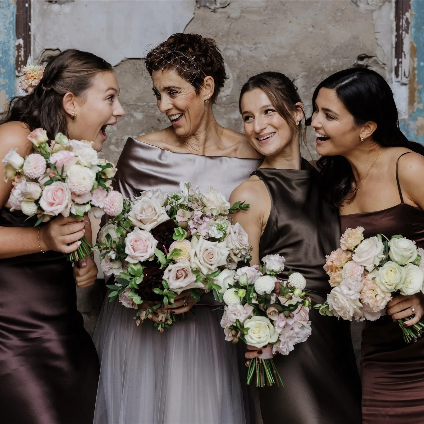 Four women at a wedding celebration, holding bouquets of pink and white flowers, dressed in dark brown satin dresses, standing close to each other and smiling.