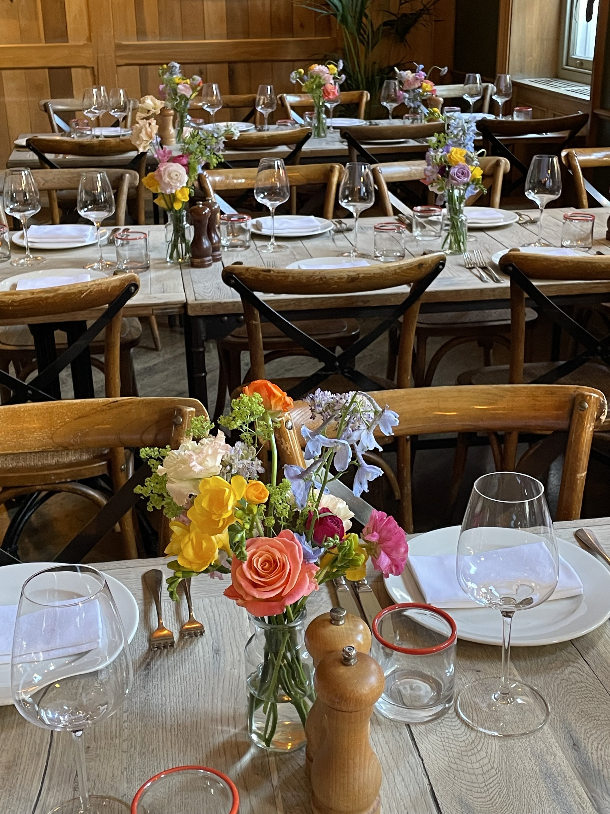 A dining table set for a meal with multiple wine glasses, white plates, silverware, small candles, and colorful floral centerpieces on a wooden table in a cozy wooden-paneled room.