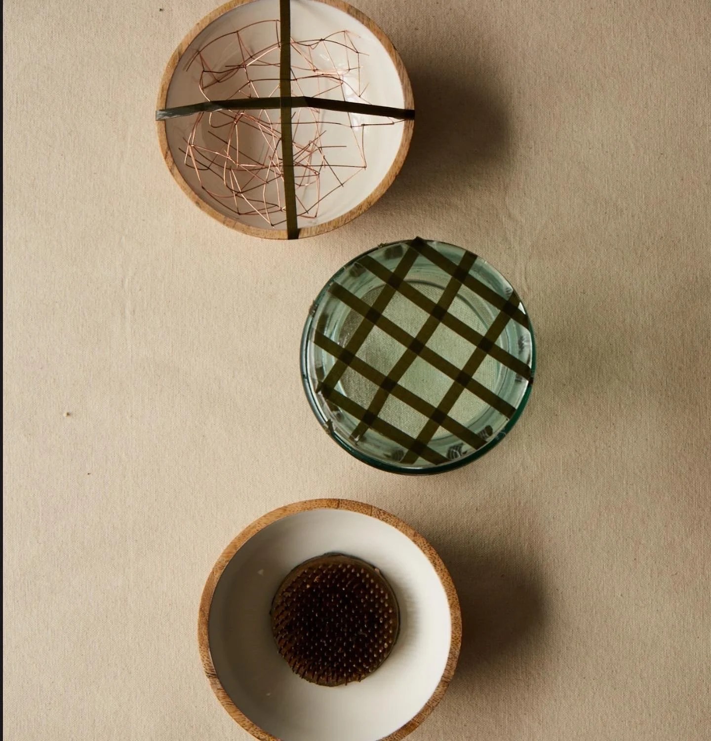 Three bowls on a beige surface, one with a wooden exterior and white interior with a round brown textured object inside, one with a glass exterior with a greenish tint and a grid pattern on its surface, and the third with a wooden exterior and white 