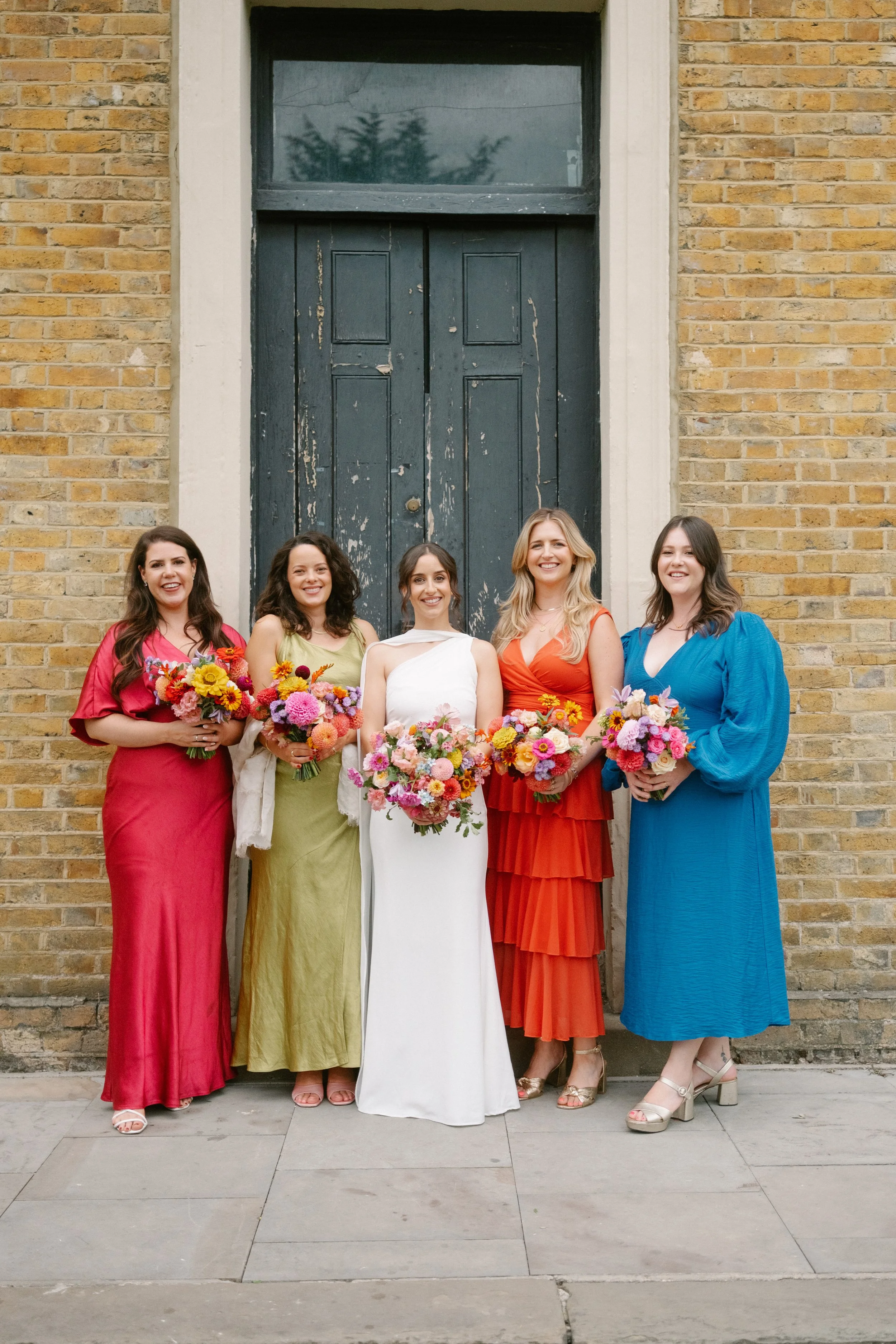 Five women standing in front of a brick building with a large dark green wooden door, each holding a colorful bouquet of flowers and dressed in bright, elegant dresses.