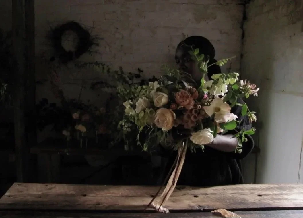 Catherine Foxwell holds a large bouquet of flowers in a dimly lit room with a rustic brick wall.