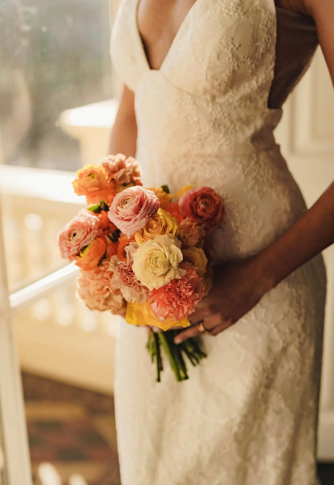 Close-up of a bride in a lace wedding dress holding a bouquet of pink, peach, and yellow flowers indoors.