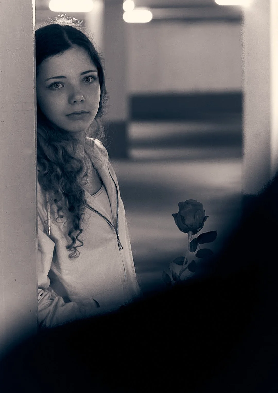A young woman with curly hair and light-colored eyes standing behind a wall, holding a rose, in a grayscale photo.