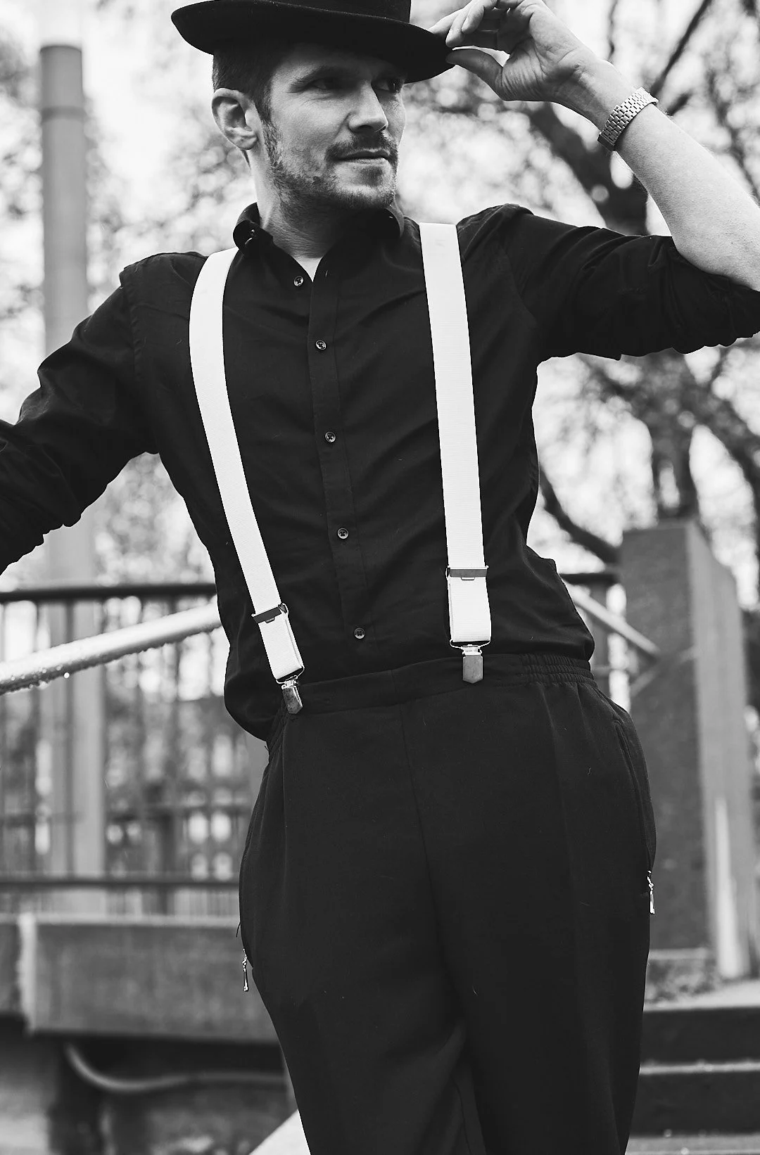 Black and white photo of a man wearing a fedora hat, suspenders, and a dress shirt, standing outdoors with trees in the background.