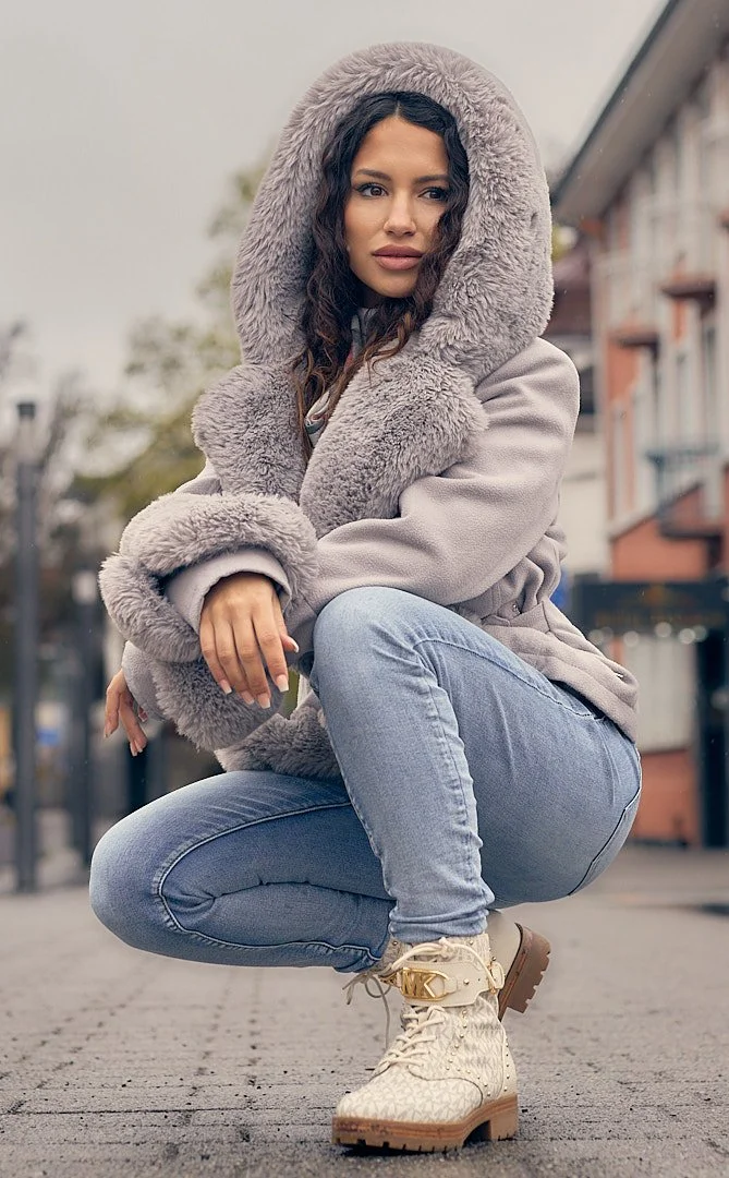 A woman crouching outdoors in a city street, wearing a gray fur-lined hooded coat, light blue jeans, and beige combat boots, with buildings and trees in the background.
