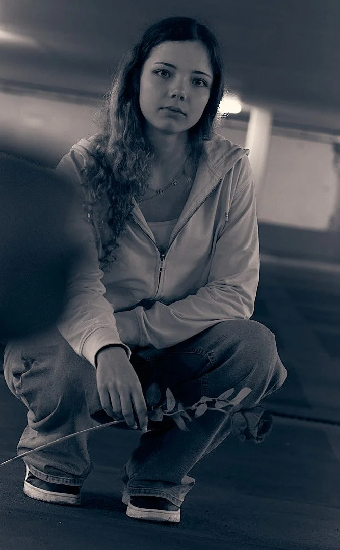 A young woman with long wavy hair, wearing a zip-up hoodie and jeans, squatting on the ground, holding a small branch, in a dim underground parking garage.