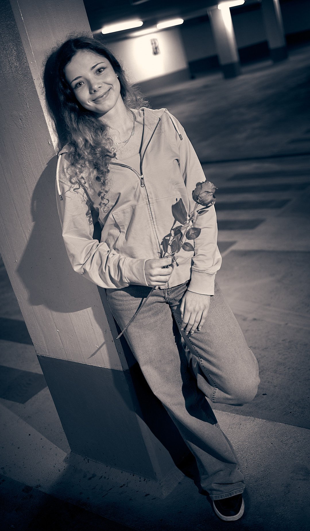 A young woman with curly hair leaning against a wall in a parking garage, holding a rose, smiling at the camera.