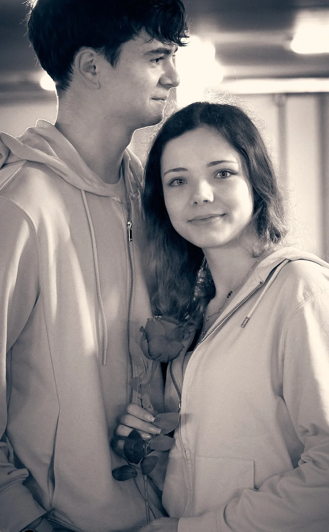 A young man and woman standing close together indoors, with the woman holding a single rose. Both are wearing light-colored zip-up hoodies, and the woman has a gentle smile. The photo is in black and white.