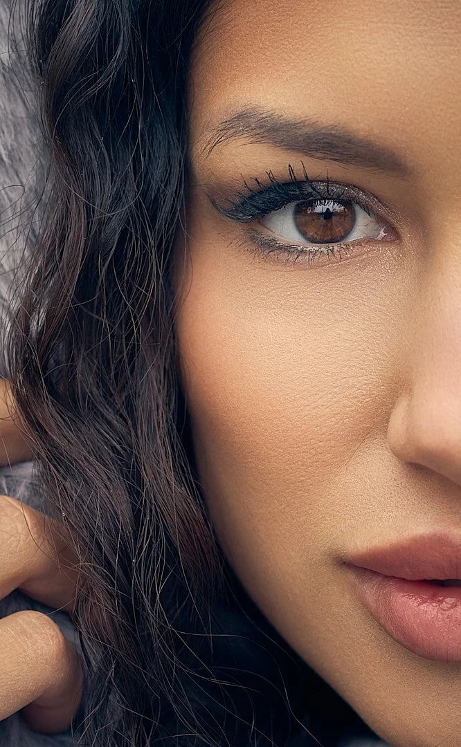 Close-up of a woman's face showing her brown eye, dark wavy hair, makeup, and part of her lips