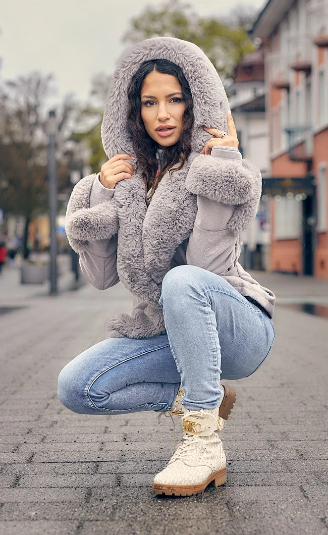 A young woman wearing a gray faux fur-lined hooded jacket, light blue jeans, and beige lace-up boots squatting outdoors on a city street with buildings and trees in the background.