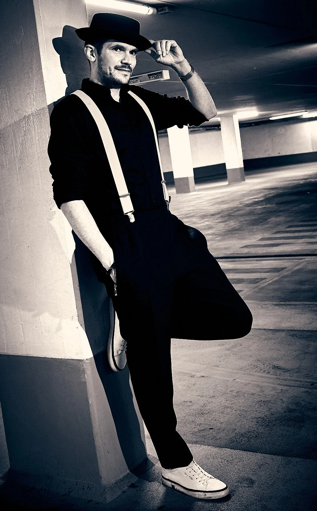 A man in a black outfit with suspenders and a hat is leaning against a parking garage wall, holding his hat with one hand and one foot crossed over the other, in a black-and-white photo.