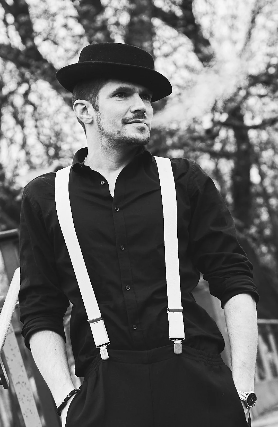 A man in a black shirt with rolled-up sleeves, suspenders, and a hat, standing outdoors with trees blurred in the background, in black and white.