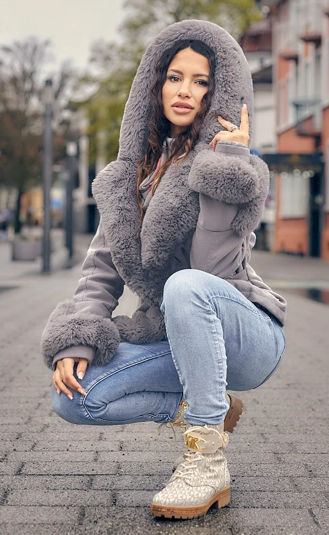 A woman squatting outdoors on a city street, wearing a large gray hooded faux fur coat, light blue jeans, and beige textured boots, with trees and buildings in the background.