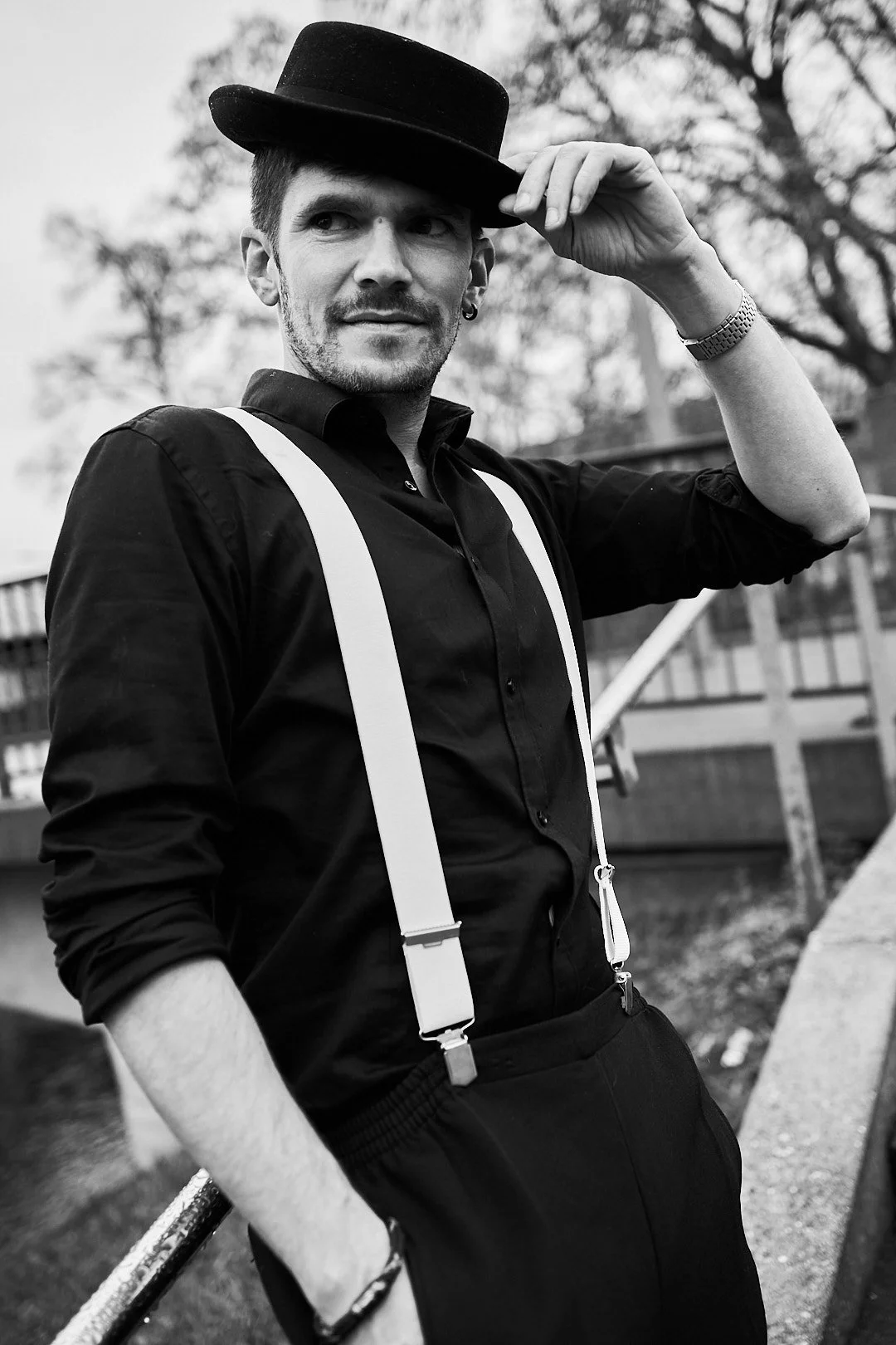 A man in a black shirt with suspenders and a hat, adjusting his hat with one hand, outdoors with trees in the background, in black and white.