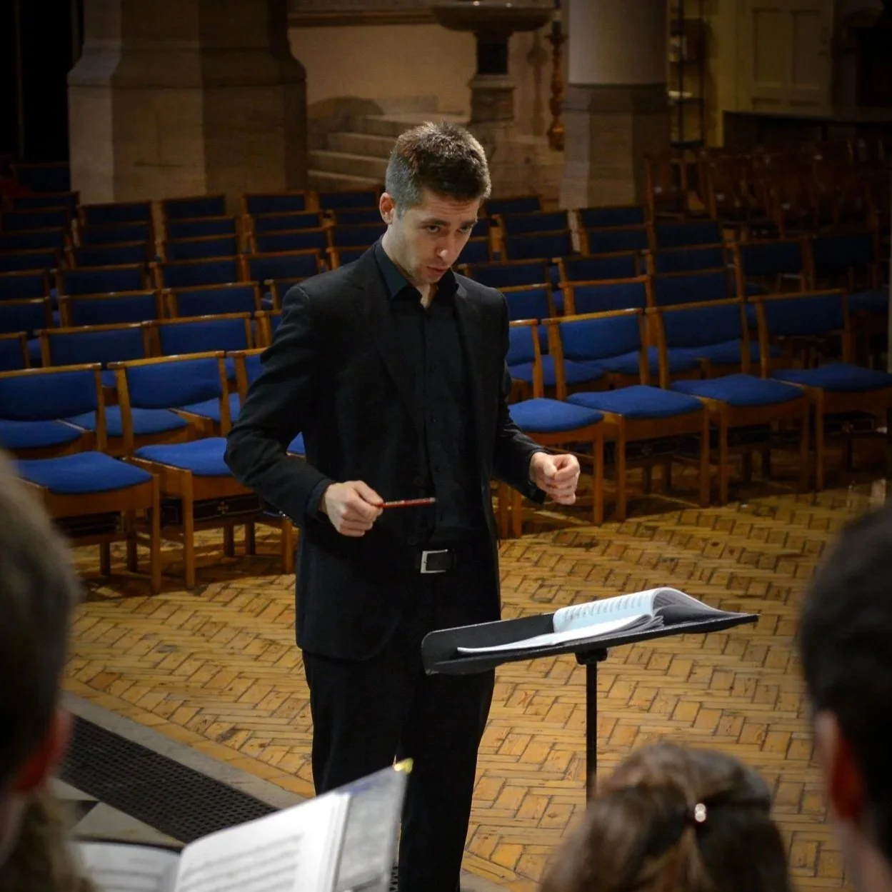 A young man in a black suit conducting an orchestra or choir, standing at a music stand with open sheet music, in a large room with wooden flooring and rows of empty blue-cushioned chairs.