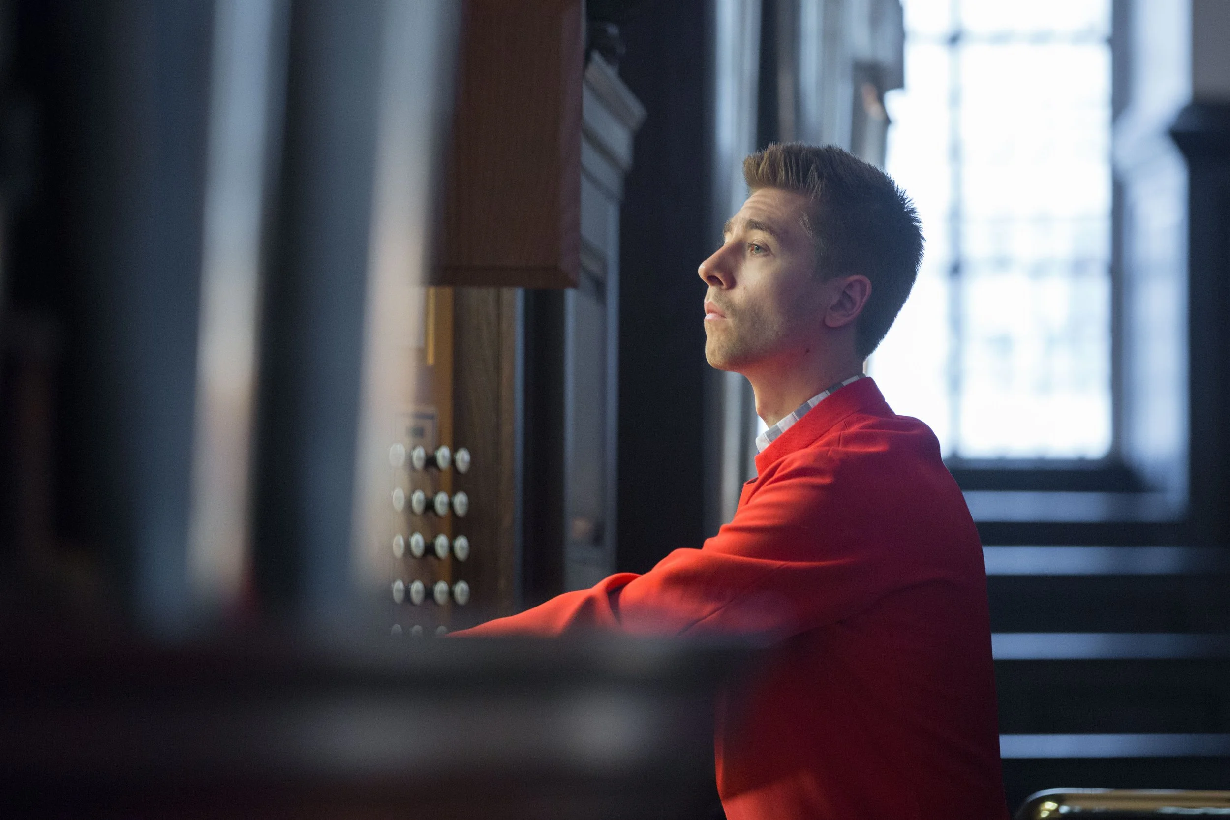 A man in a red jacket sitting at an organ in a church or cathedral, with natural light coming through large windows in the background.