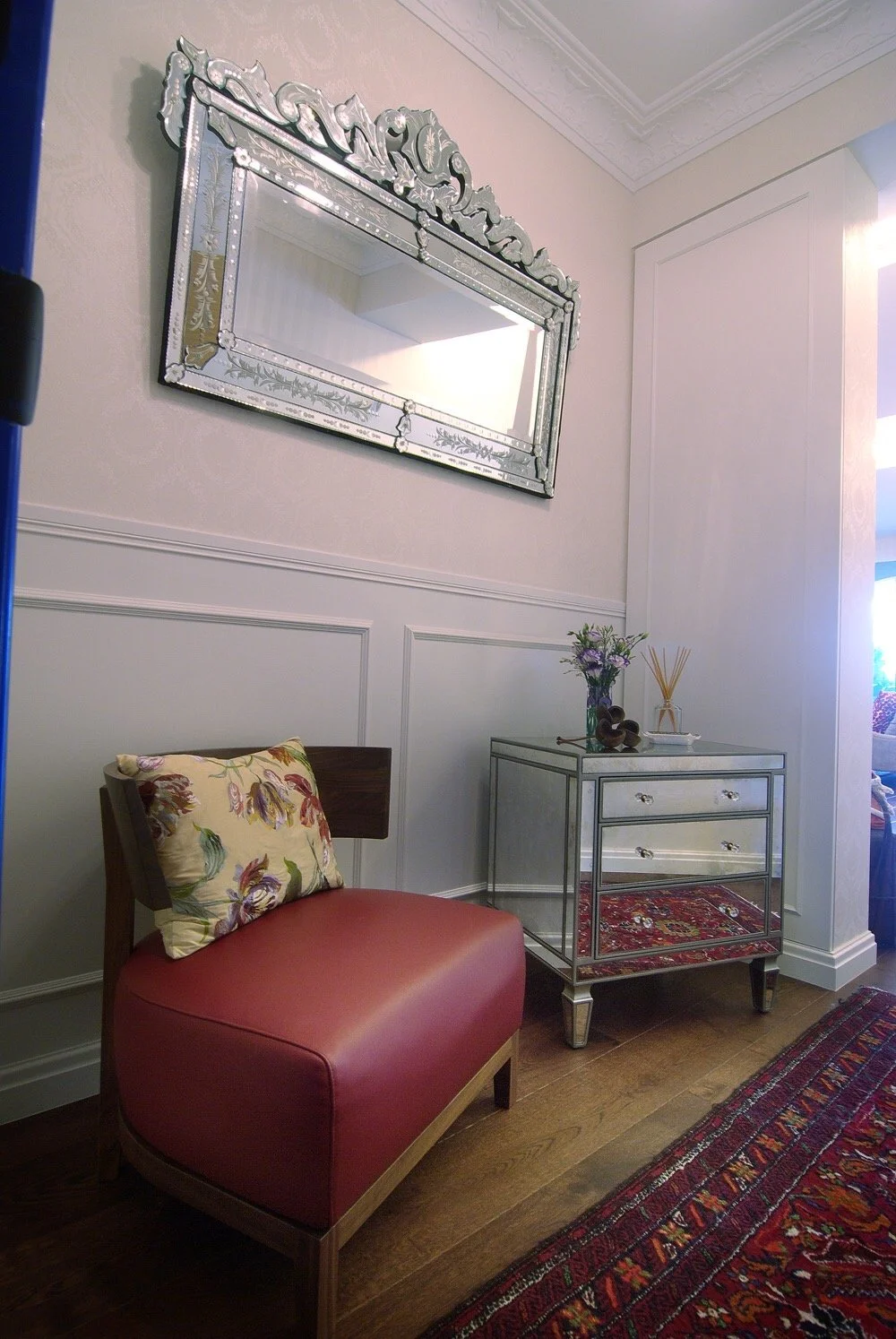 A corner of a room with a red upholstered chair with a floral cushion, a mirrored silver side table with drawers, a glass vase with purple flowers, a reed diffuser, and a decorative mirror hanging on the wall. There's a colorful patterned rug on the wooden floor.