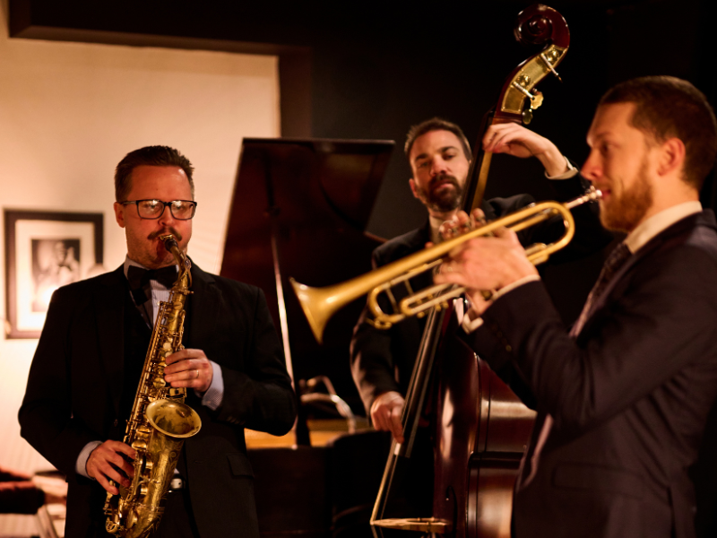 Three male jazz musicians performing with saxophone, double bass, and trumpet.