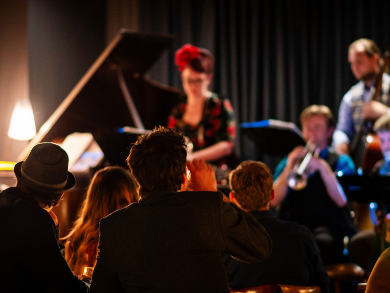 A jazz band performs live with a pianist, bassist, and trumpeter, while an audience watches in a dimly lit venue.