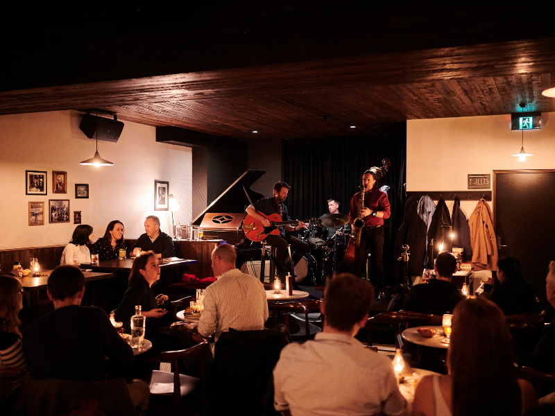 Live jazz band performing on stage at a cozy nightclub with audience seated at tables, intimate lighting, and dark curtains in the background.