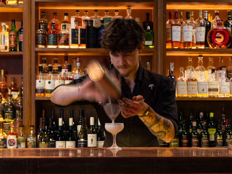Bartender pouring a cocktail into a martini glass at a bar with shelves of liquor bottles in the background.