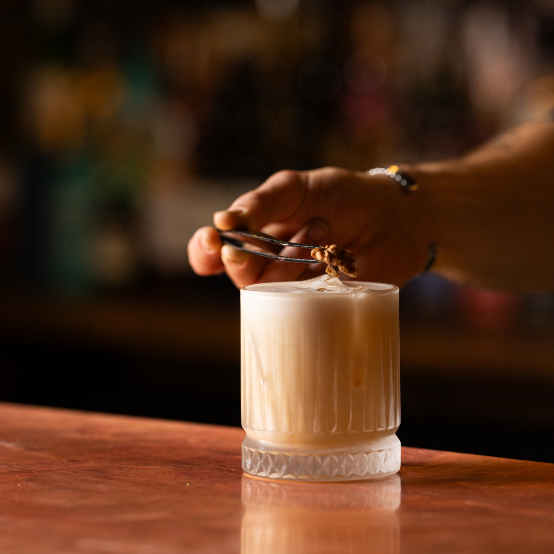 A hand garnishing a creamy cocktail with a small element using tweezers, set on a wooden bar counter with a blurred background.