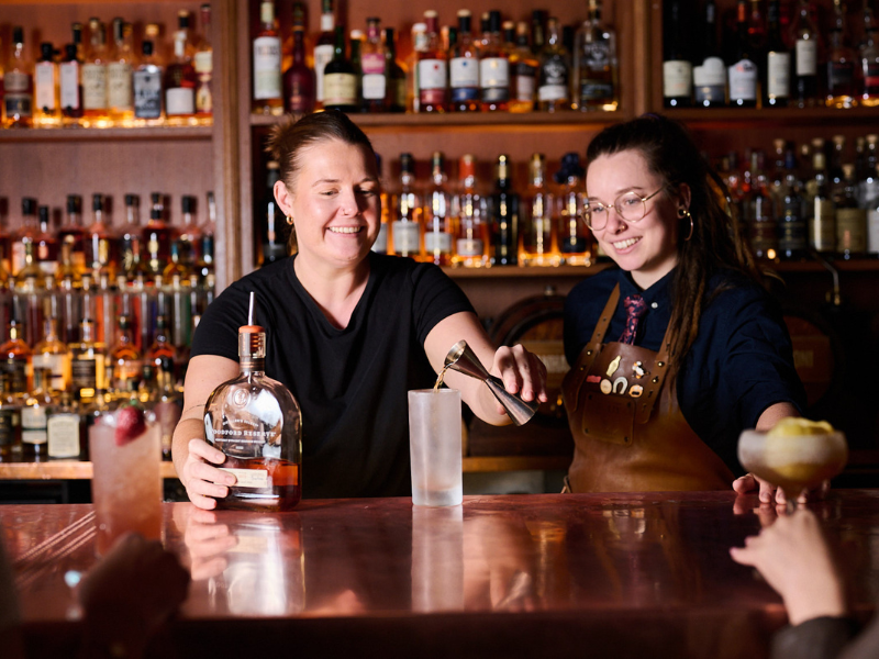 Two women at a bar preparing drinks, with a variety of liquor bottles on shelves in the background. One woman is pouring a drink into a glass while the other watches and smiles, holding a bowl.