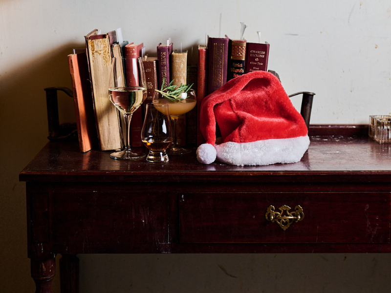 A wooden table with books, two glasses of wine, a sprig of herbs, and a red Santa hat with white trim and pom-pom, against a white wall.