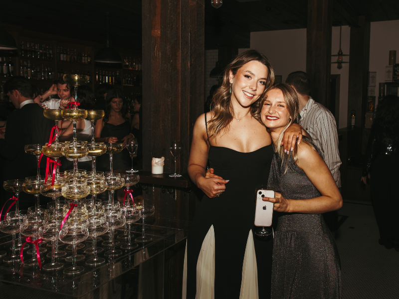 Two women smiling and hugging at a celebration, standing next to a pyramid of champagne glasses decorated with pink ribbons in a dimly lit party setting.