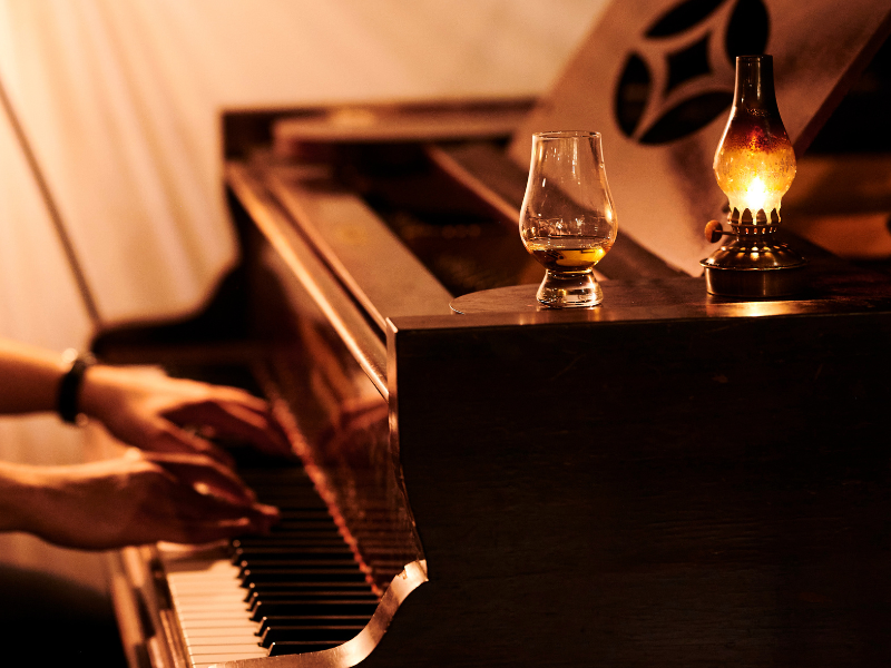 A person playing a piano at a cozy, warmly lit setting with a glass of whiskey and a decorative lamp on the piano