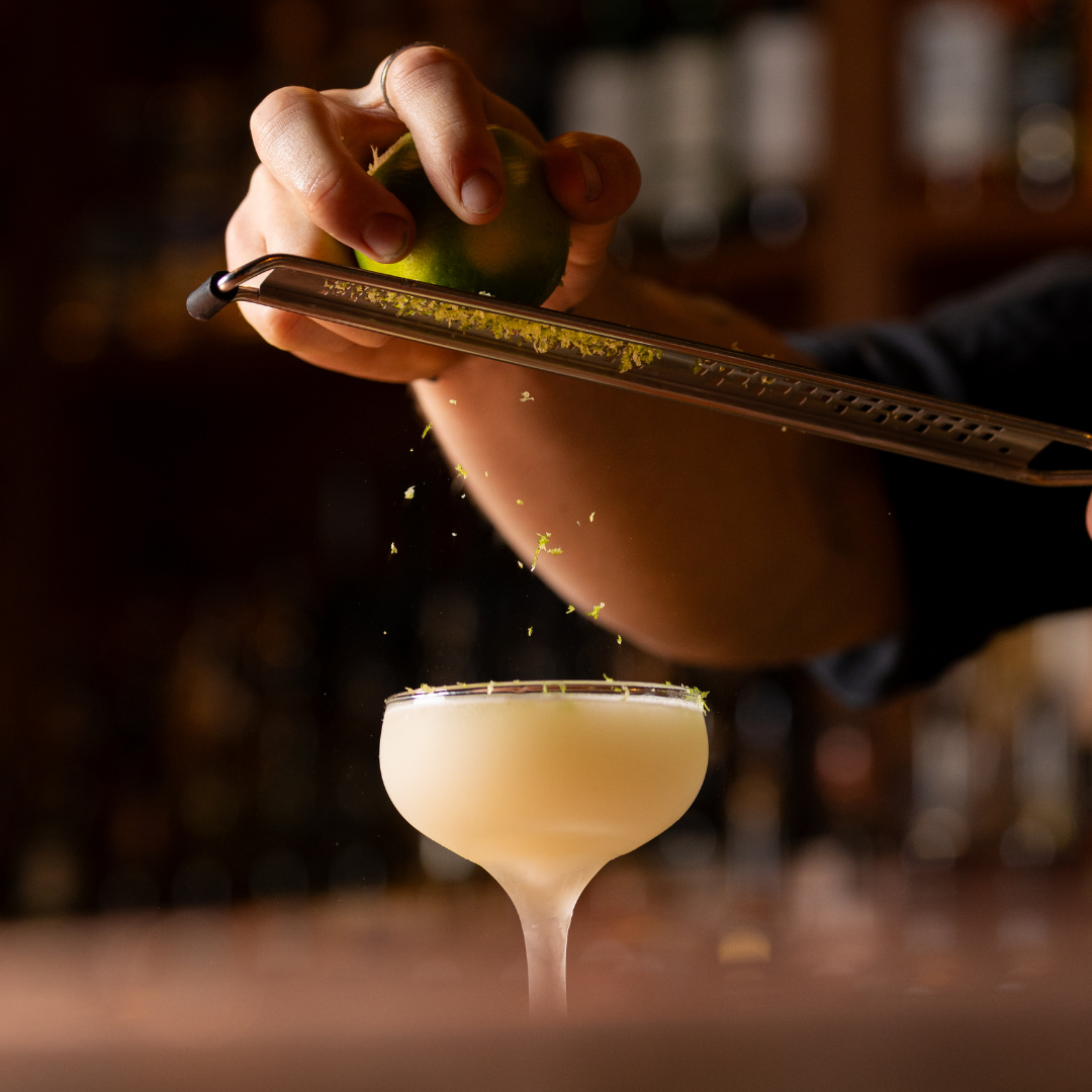 Person grating lime peel into a cocktail glass